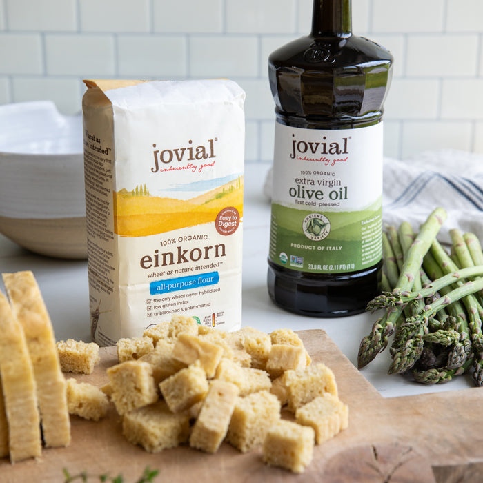 Herby cubed einkorn croutons on a cutting board with Jovial einkorn flour bag, extra virgin olive oil bottle, and asparagus