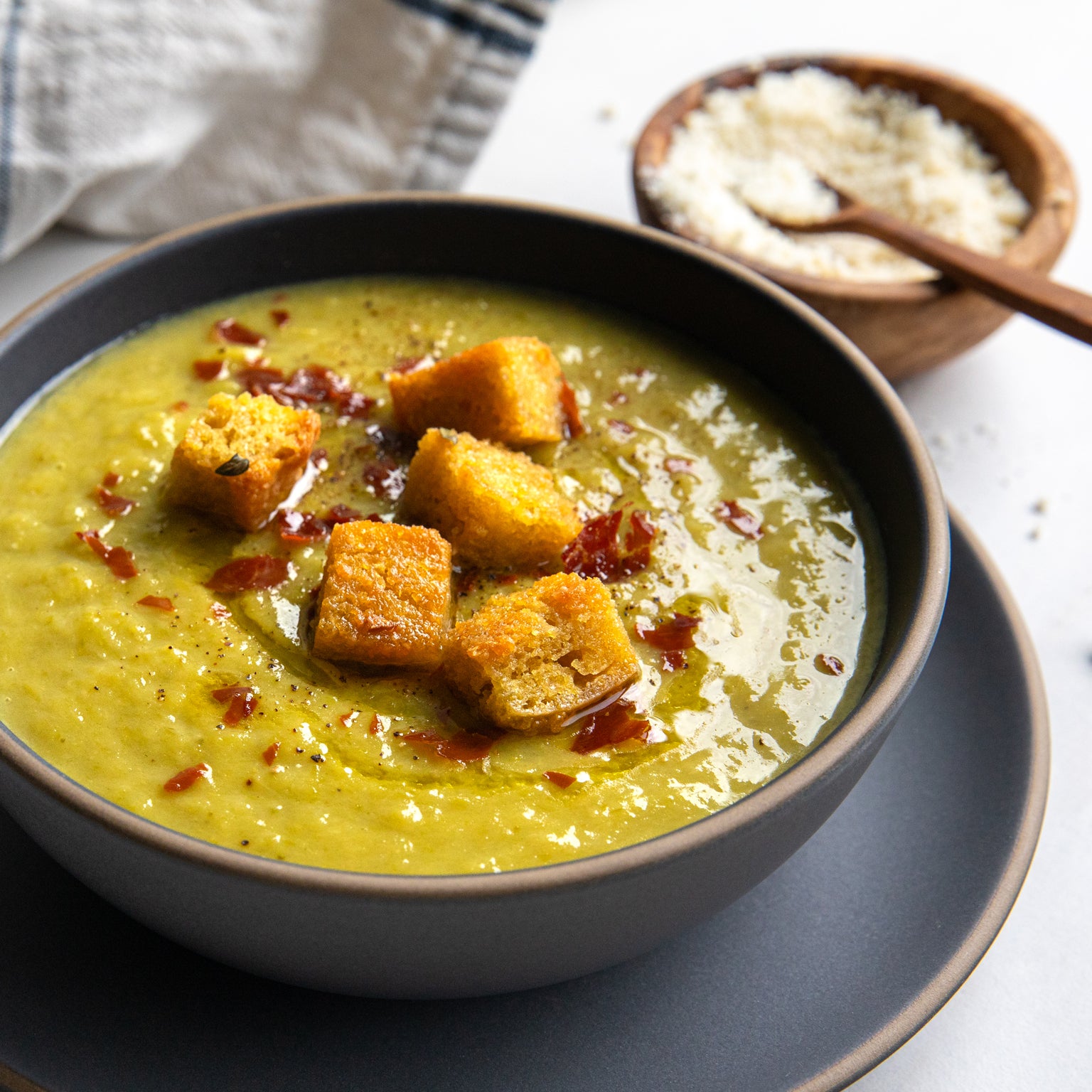Bowl of asparagus vellutata topped with golden herby einkorn croutons and chili flakes on a gray saucer