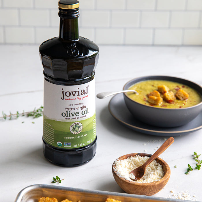 Jovial extra virgin olive oil bottle beside a bowl of asparagus soup topped with herbed einkorn croutons, a wooden bowl of grated cheese, and a tray of croutons on white marble