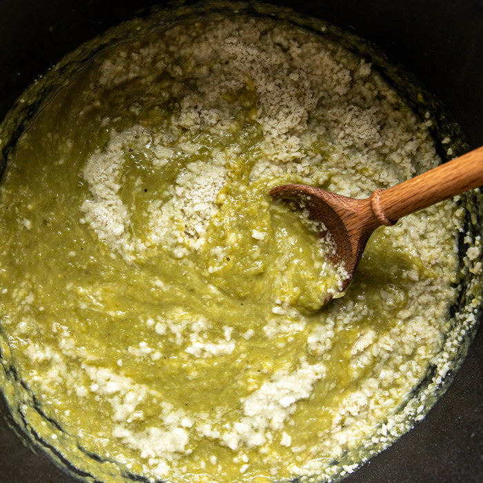 Asparagus soup being stirred with a wooden spoon in a dark pot, topped with crumbled herby einkorn croutons