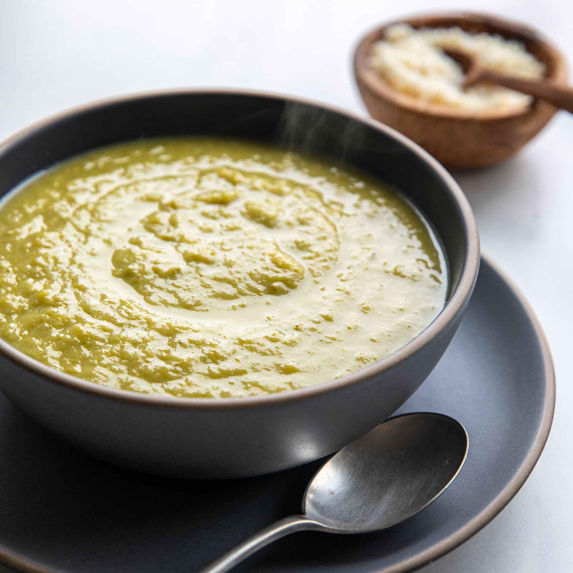 Hot velvety asparagus soup served in a gray bowl with a spoon on a matching saucer and a small bowl of grated cheese in the background