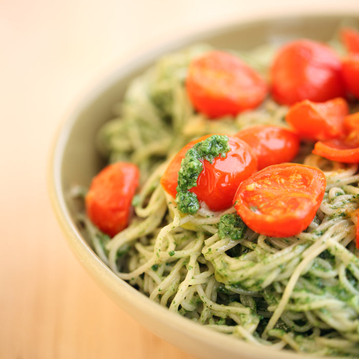 Close-up of bowl of capellini tossed in spinach pesto and topped with roasted cherry tomatoes on a light wood background