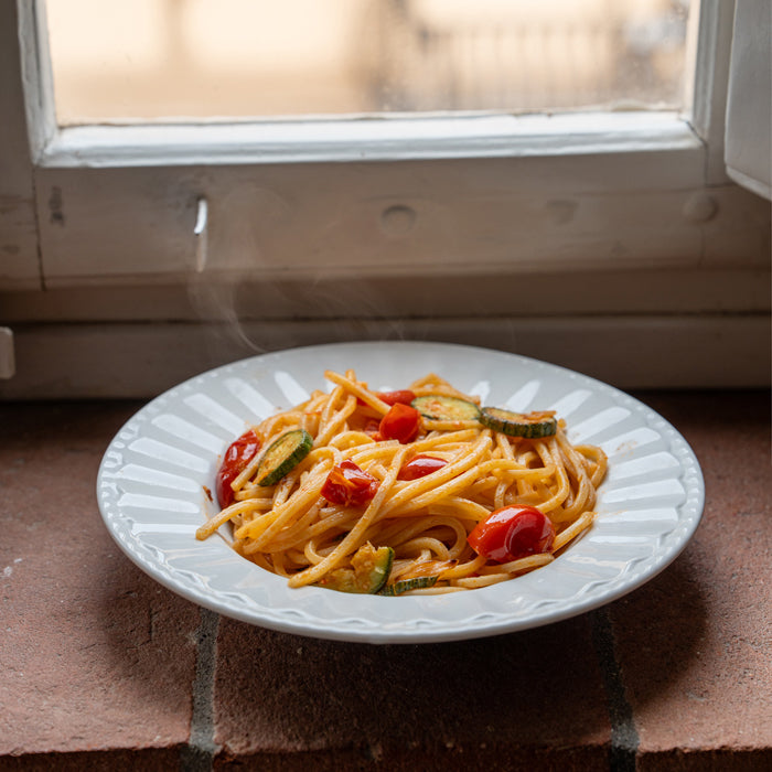 Steaming plate of spaghetti with zucchini, roasted cherry tomatoes, and basil on a white plate set on a windowsill