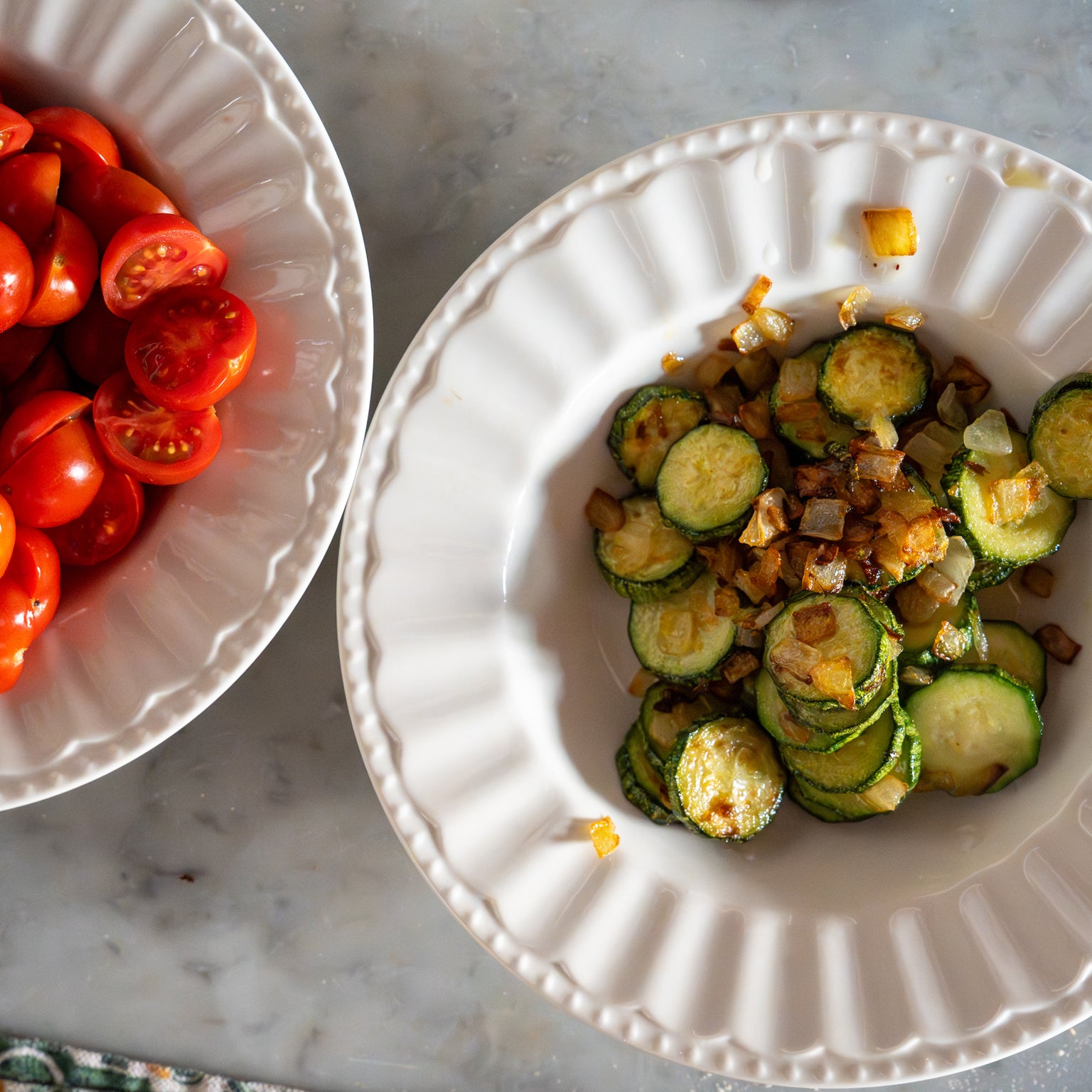 Overhead view of two white bowls on marble: halved cherry tomatoes in one and sautéed zucchini with caramelized onions in the other