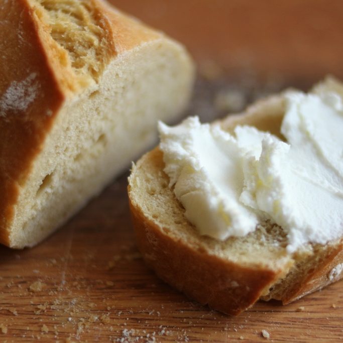 Close-up of sliced einkorn baguette showing a slice spread with soft white cheese on a wooden cutting board