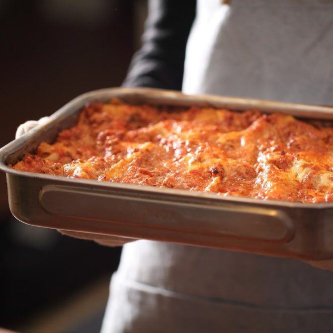 Gluten-free lasagna Bolognese being held in a metal baking pan by a person wearing an apron