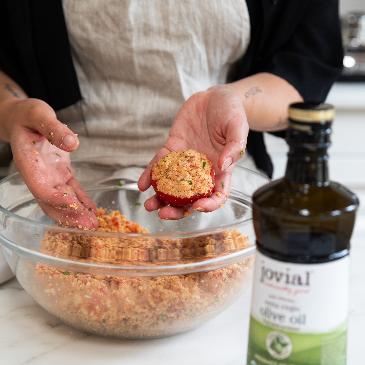 Hands stuffing a halved tomato with einkorn breadcrumbs over a glass mixing bowl, Jovial olive oil bottle in the foreground