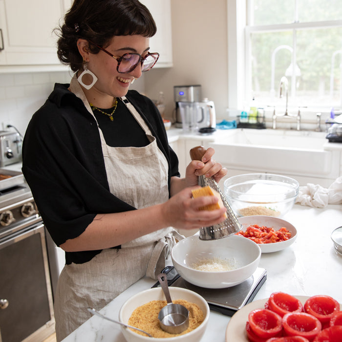 Home cook grating cheese over bowls of Einkorn breadcrumbs and diced tomatoes on a kitchen counter