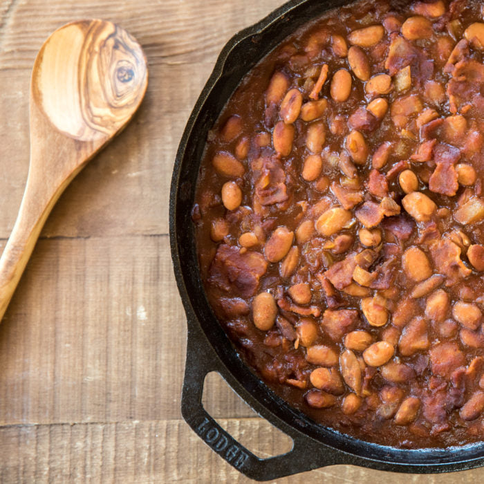 Top-down view of baked borlotti beans with bacon and tomato sauce in a cast-iron skillet on a wooden table, wooden spoon at left