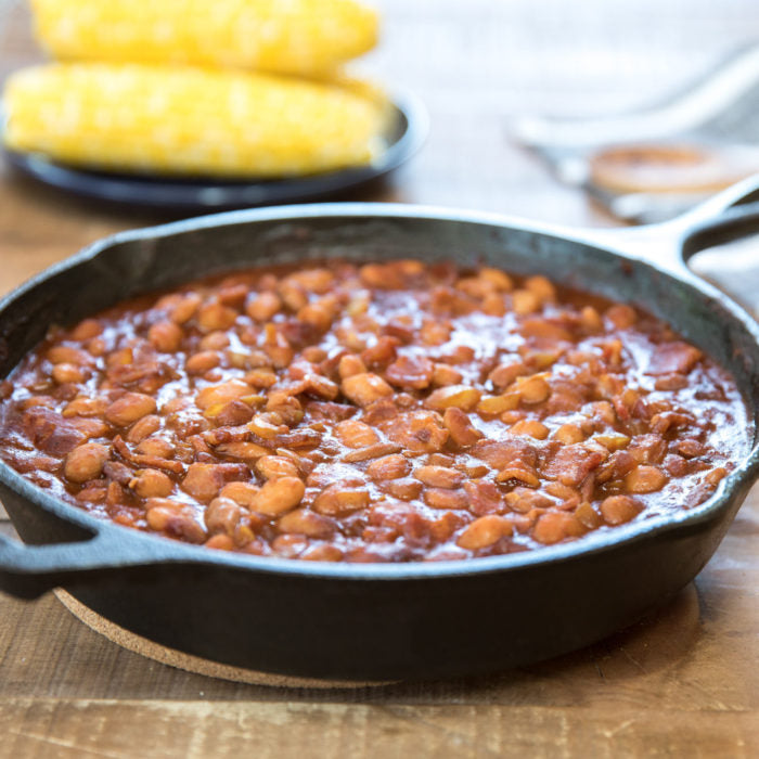 Cast-iron skillet of organic borlotti beans in tomato-bacon sauce with blurred corn in background