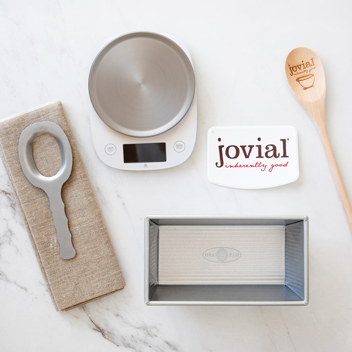 Top view of Baker’s Tool Kit: scale, loaf pan, linen couche, kneading tool, Jovial scraper, wooden spoon on light marble