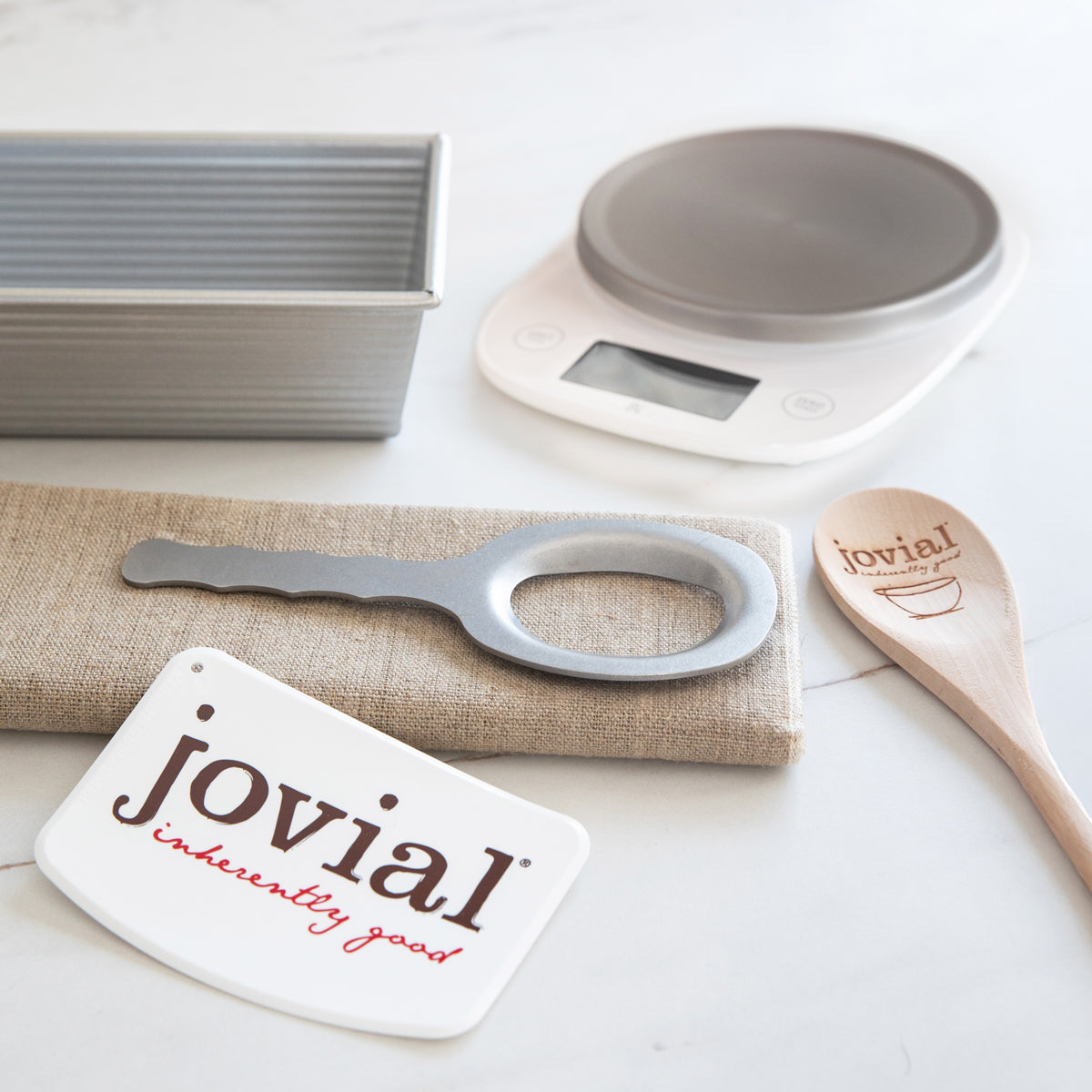 Angled view of Baker’s Tool Kit showcasing loaf pan, baking scale, wooden spoon, dough scraper, and linen couche on white