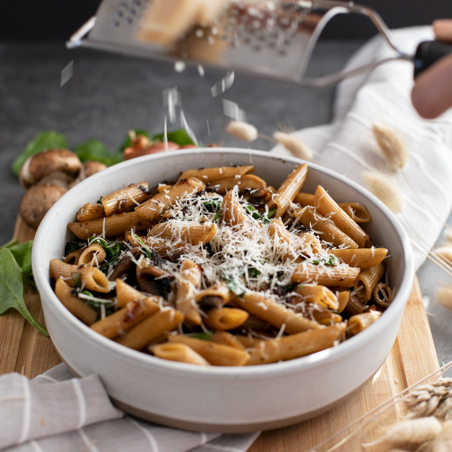 Bowl of cooked organic whole wheat penne rigate with mushrooms, spinach, and grated cheese on a wooden board