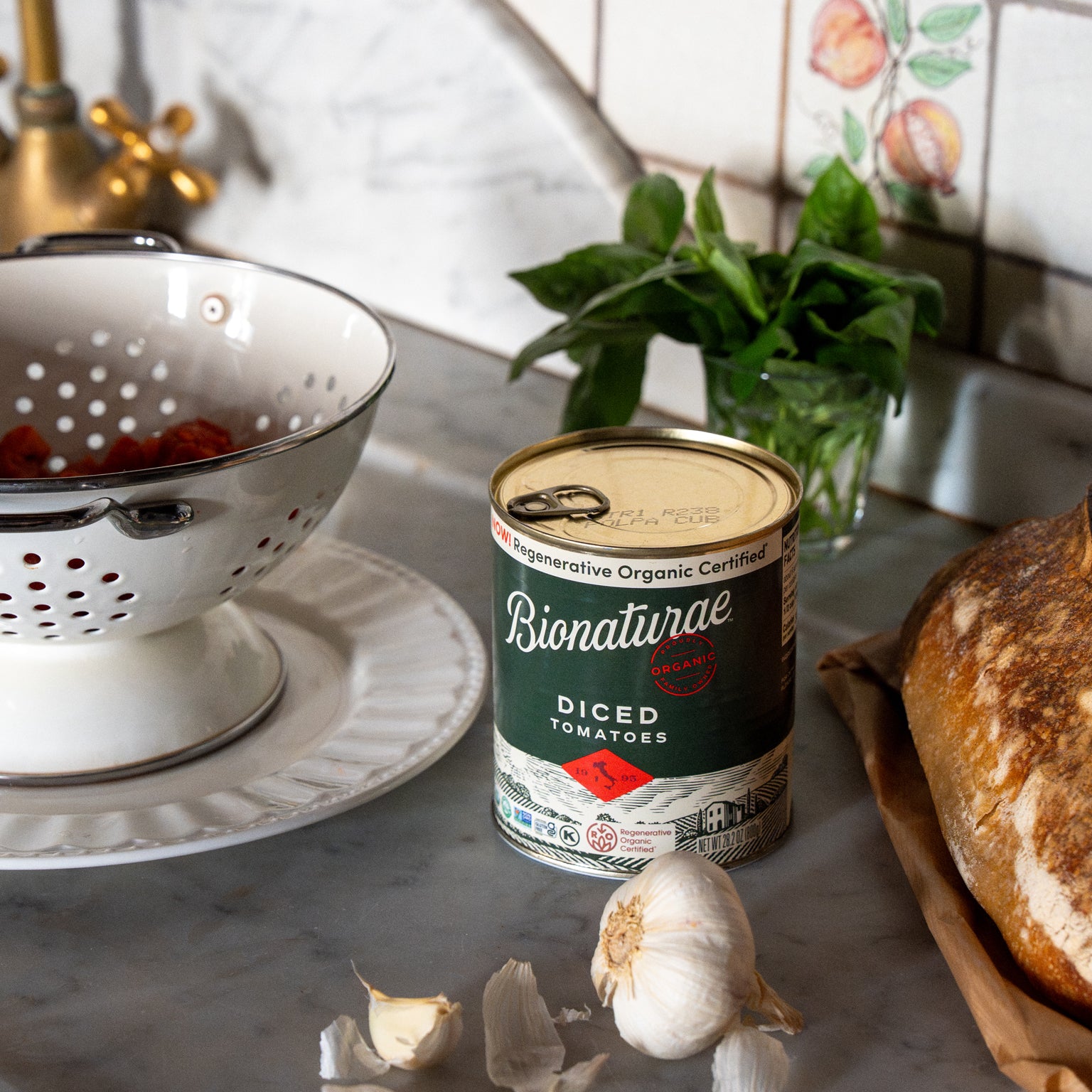 Can of diced tomatoes on a marble counter beside a colander of tomatoes, garlic cloves, fresh basil, and crusty bread