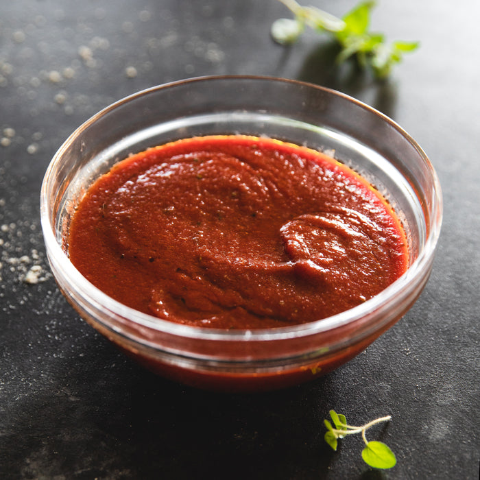 Overhead view of organic Bionaturae strained tomatoes pizza sauce in a glass bowl showing smooth thick texture on dark surface with oregano sprig