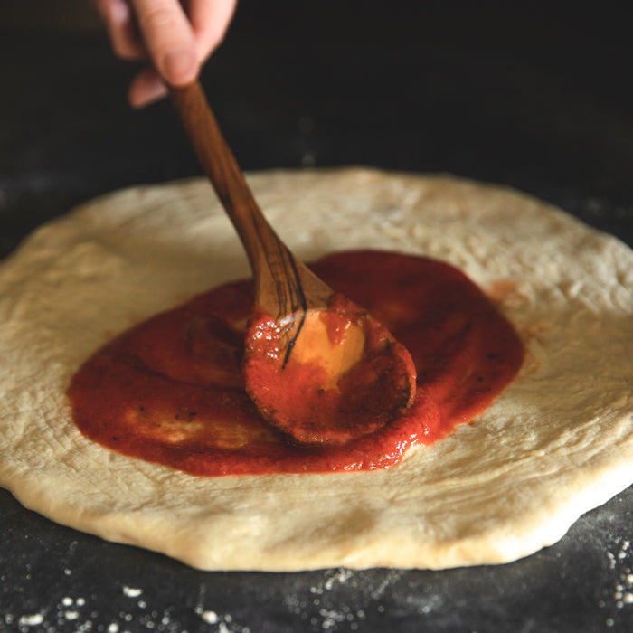 Hand spreading Bionaturae strained tomato sauce with a wooden spoon onto pizza dough on a dark surface