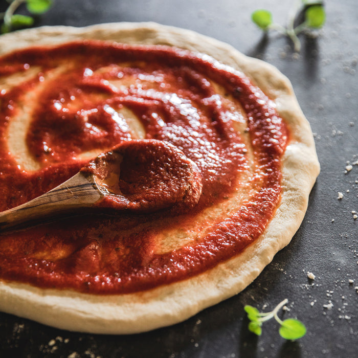 Pizza dough with Bionaturae strained tomato sauce being spread with a wooden spoon on a dark countertop