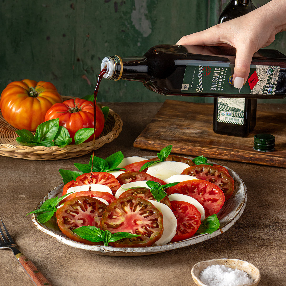 Hand pouring organic balsamic vinegar from a bottle onto a tomato, mozzarella, and basil salad