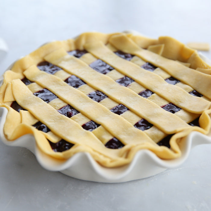 Close-up of unbaked nine-inch blueberry pie with lattice einkorn crust in a fluted white ceramic dish on a marble countertop