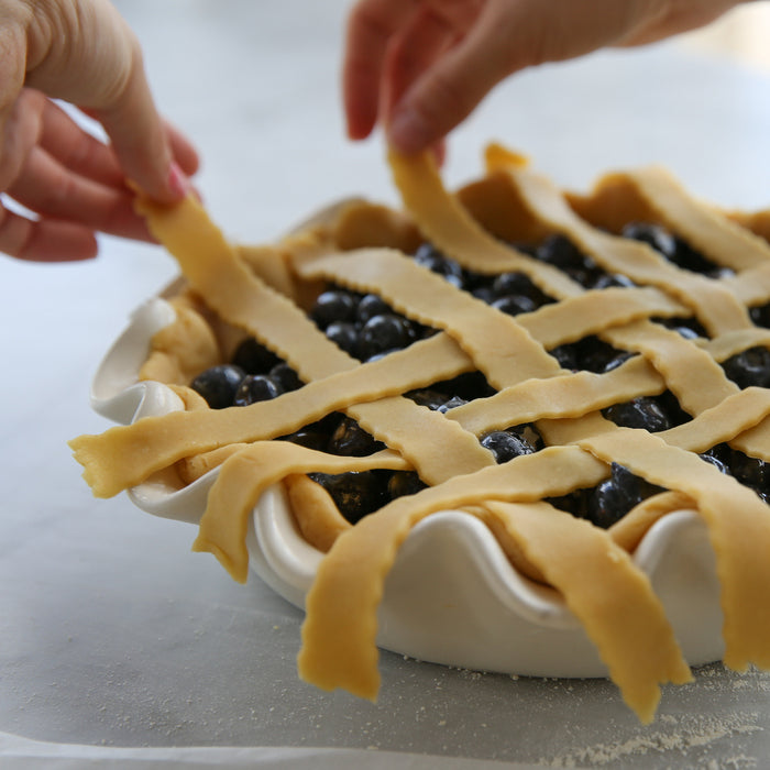 Hands weaving a lattice crust over a honey-sweetened einkorn blueberry pie in a ceramic pie dish on a marble countertop