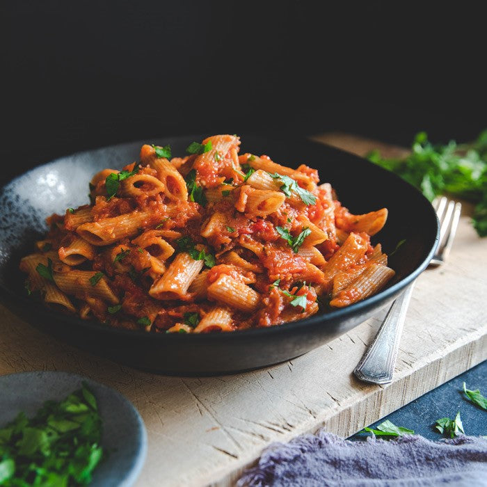 Penne with eggplant bolognese in a black bowl, whole wheat penne tossed in tomato sauce and garnished with parsley on a wooden board
