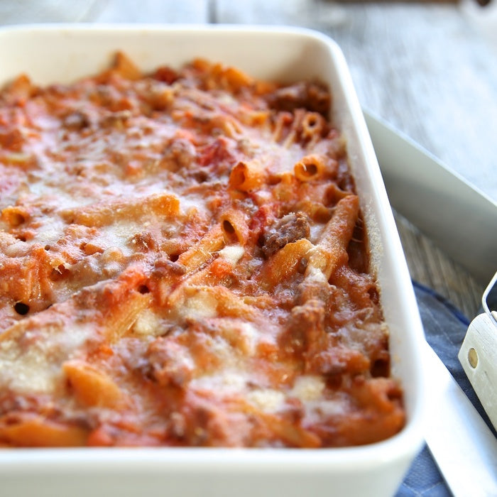 Close-up of penne Bolognese pasta bake in white baking dish showcasing melted cheese and Italian tomato sauce on wooden table