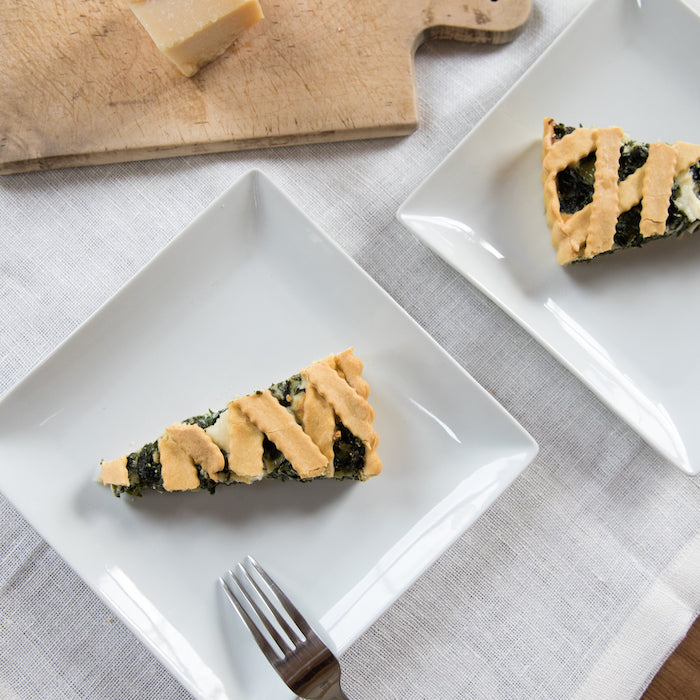 Two slices of Einkorn spinach and ricotta pie on plates with a fork and a wooden cutting board with cheese on a linen cloth