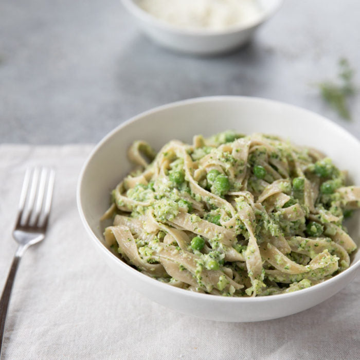 Close-up of tagliatelle pasta showcasing creamy broccoli-and-pea sauce in a white bowl on a light linen background