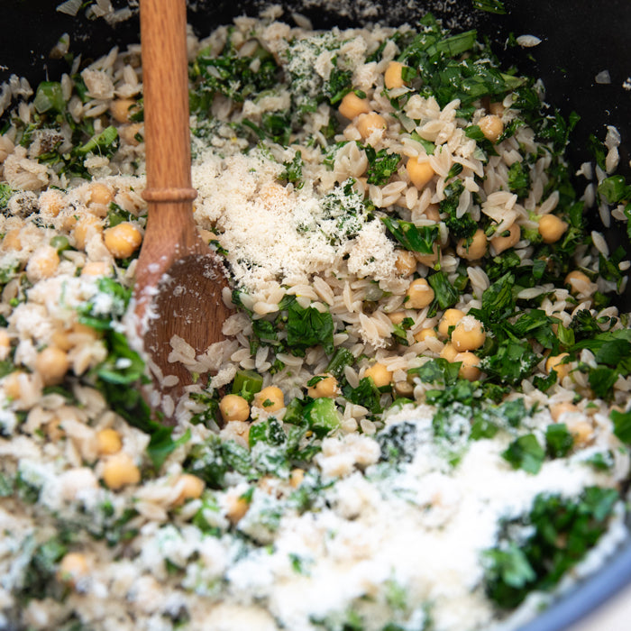 Close-up of cooked orzo with chickpeas, chopped broccolini and grated cheese being stirred in a pot with a wooden spoon