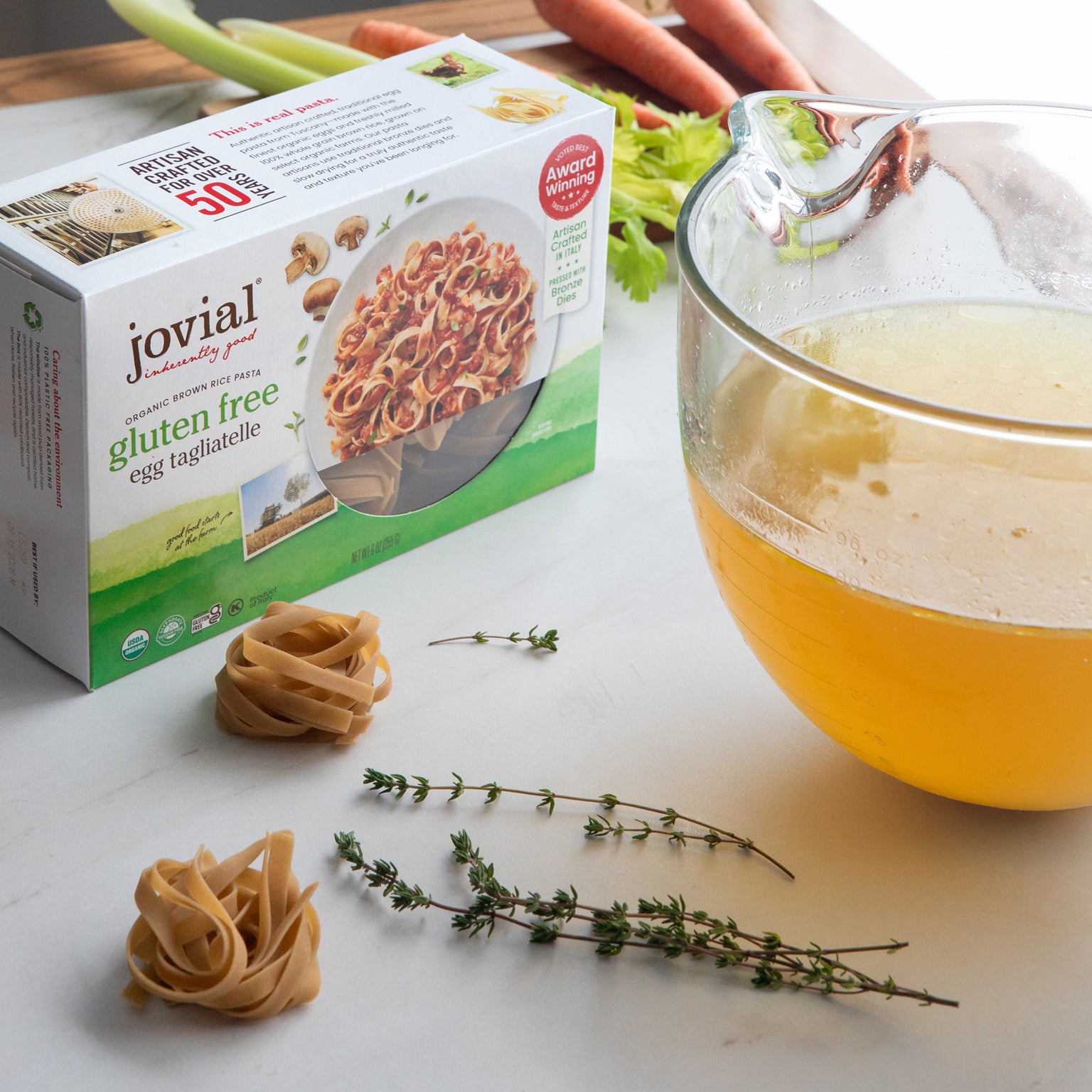 Angled view of Jovial gluten-free brown rice egg tagliatelle box showing two pasta nests, fresh thyme, and a bowl of golden broth