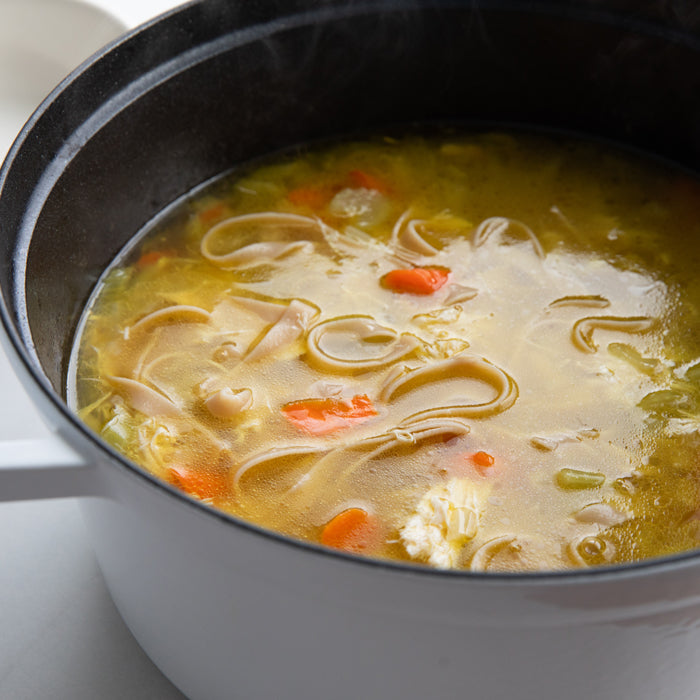 Homemade chicken noodle soup simmering in a white pot with gluten free egg tagliatelle, shredded chicken, and carrot pieces