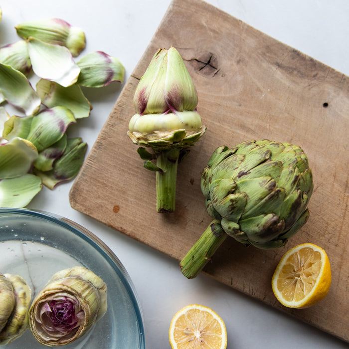 Overhead view of organic artichokes prepped on a wooden cutting board with lemon halves and a bowl of trimmed artichokes