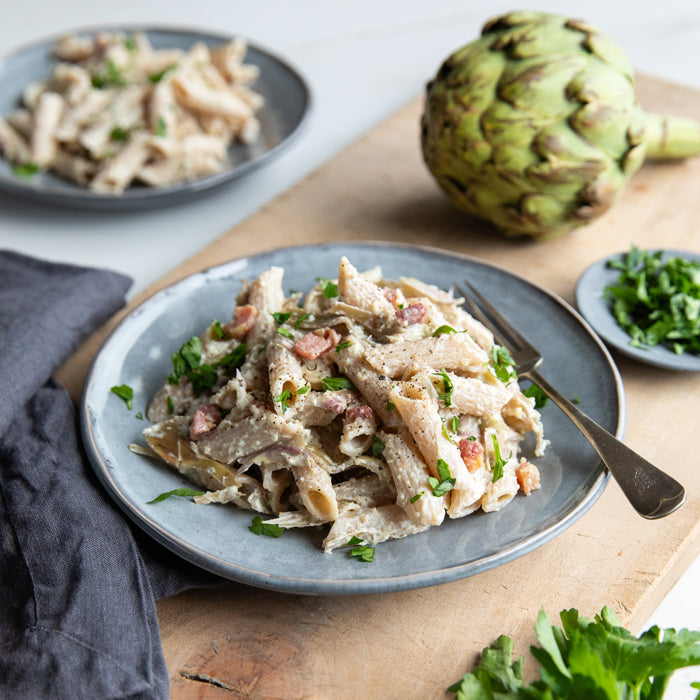 Plate of organic Brown Rice Penne tossed in creamy artichoke sauce with crispy pancetta and parsley, on a wooden board