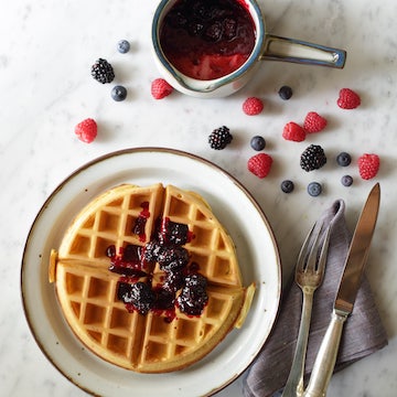 Classic Belgian waffle topped with mixed berry compote on a plate, fork and knife on a napkin, scattered berries on marble surface