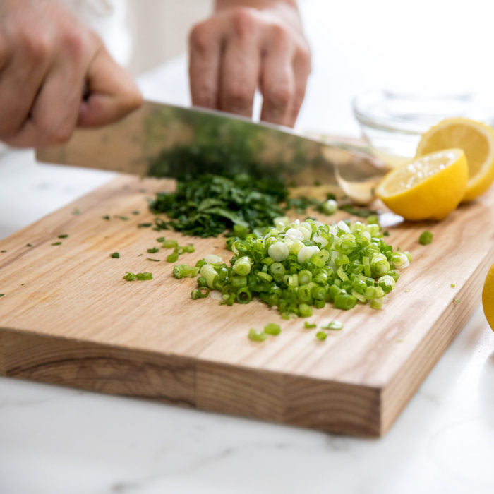 Hands chopping parsley and scallions on a wooden cutting board with lemon halves and a knife, prepping Italian bean salad