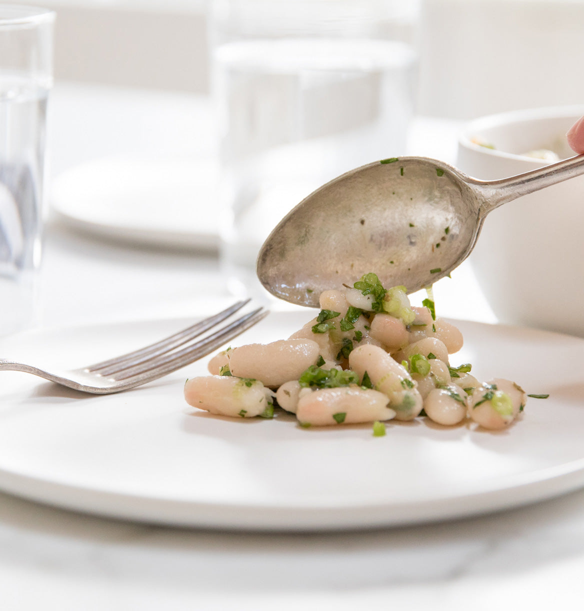 Spoon serving Italian cannellini bean salad with herbs and olive oil onto a white plate in a bright dining setting