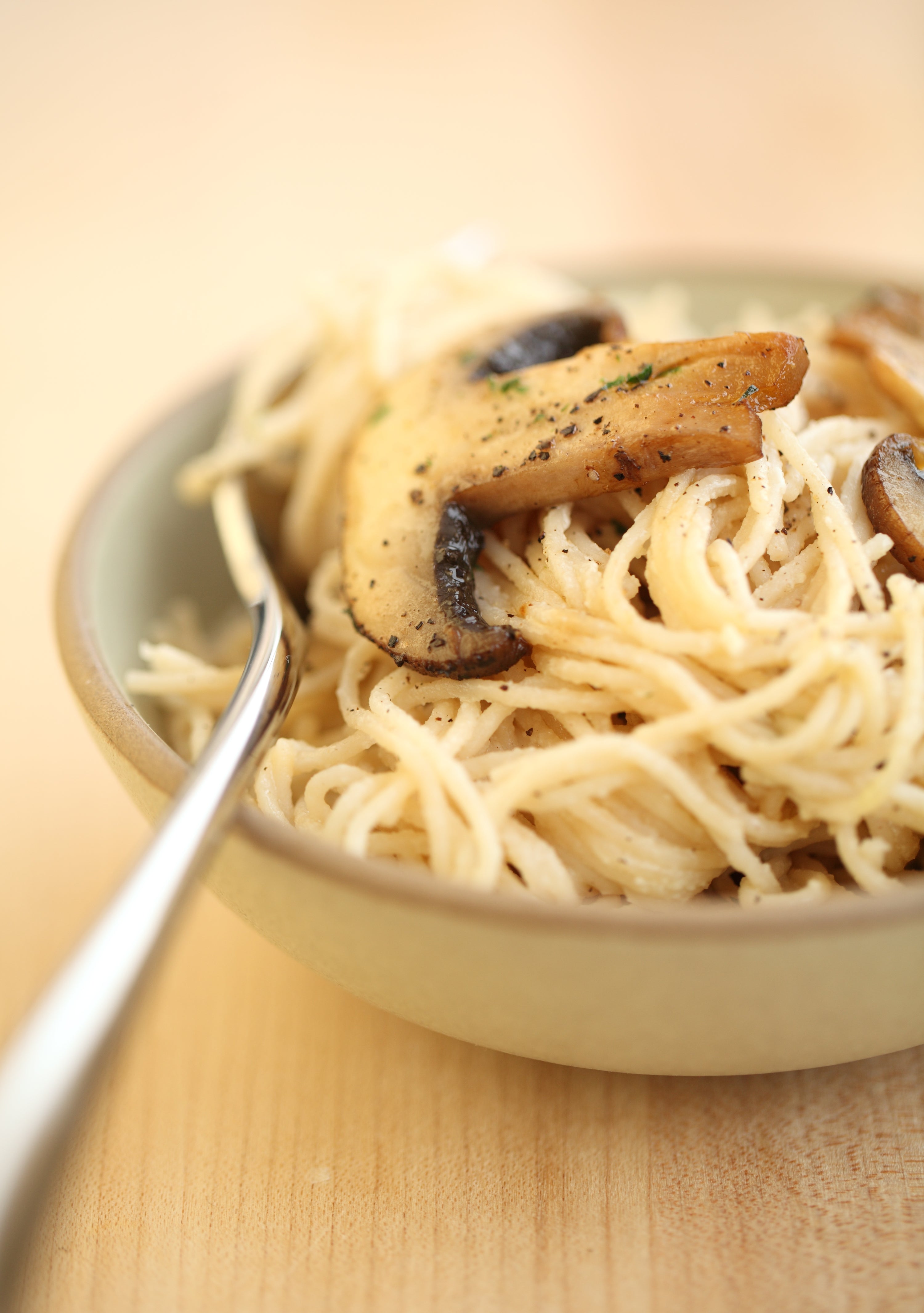 Close-up of capellini with goat cheese and pan-fried mushrooms in a beige bowl, fork at side on a light wood surface
