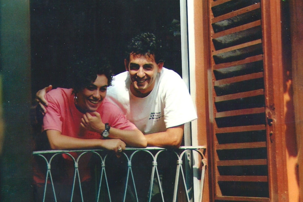 Two Jovial founders leaning on a balcony railing at a window with wooden shutters, smiling in warm sunlight
