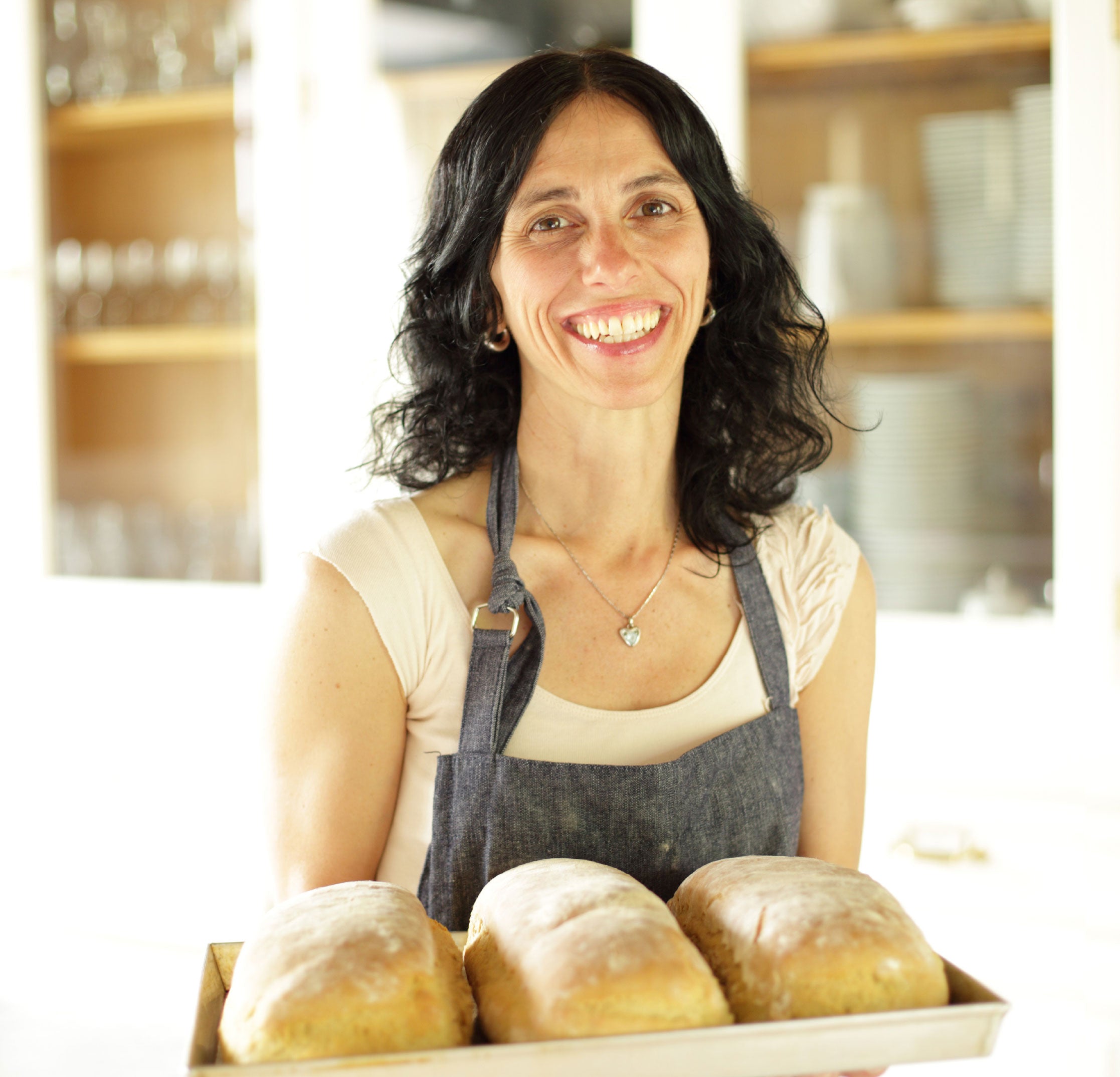 Carla, Jovial Foods co-founder, smiling and holding a tray of three freshly baked loaves in a bright kitchen