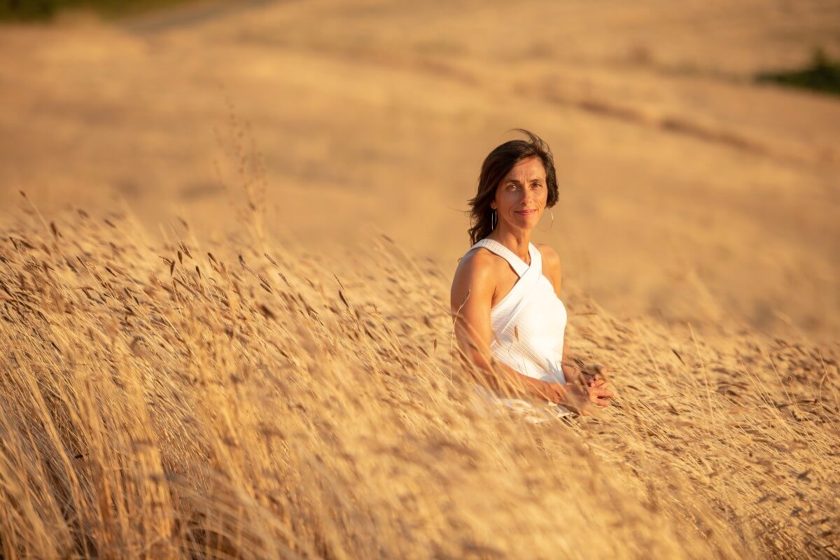 Woman standing in a golden einkorn wheat field at sunset, wearing a white sleeveless dress and looking toward the camera