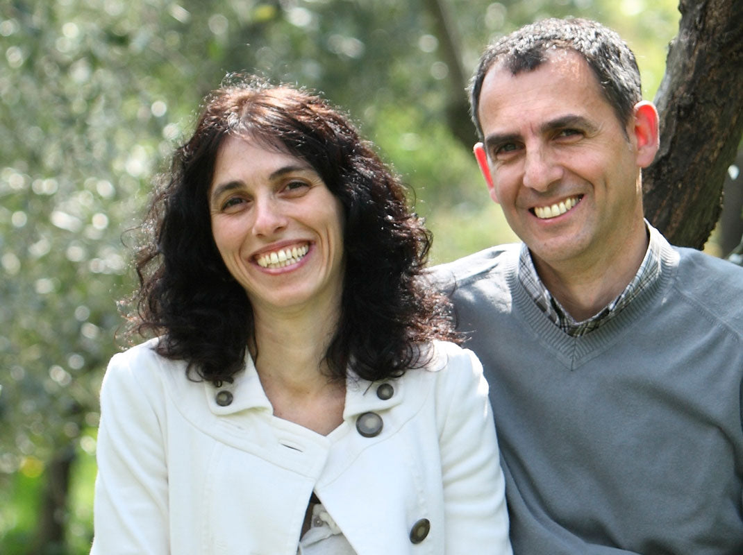 Jovial Foods founders smiling outdoors among olive trees, woman in a white jacket and man in a gray sweater