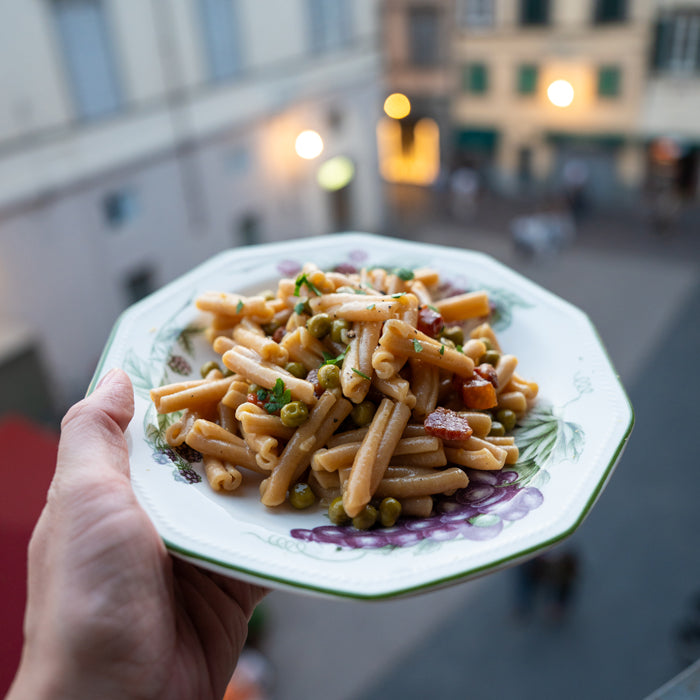 Hand holding a plate of Caserecce Carbonara with peas and pancetta, blurred outdoor Italian street in background