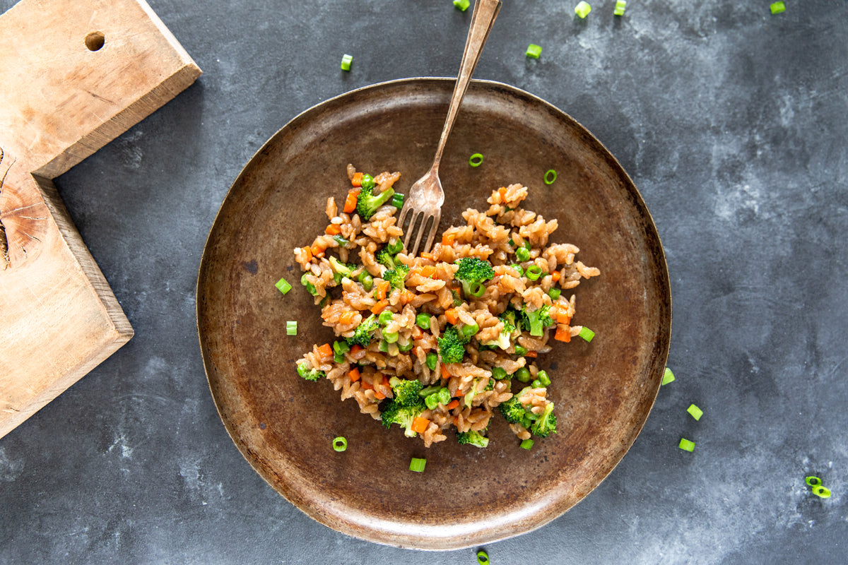 Gluten-free cassava orzo fried rice served in a brown bowl with broccoli, carrots, peas, and scallions on a dark tabletop