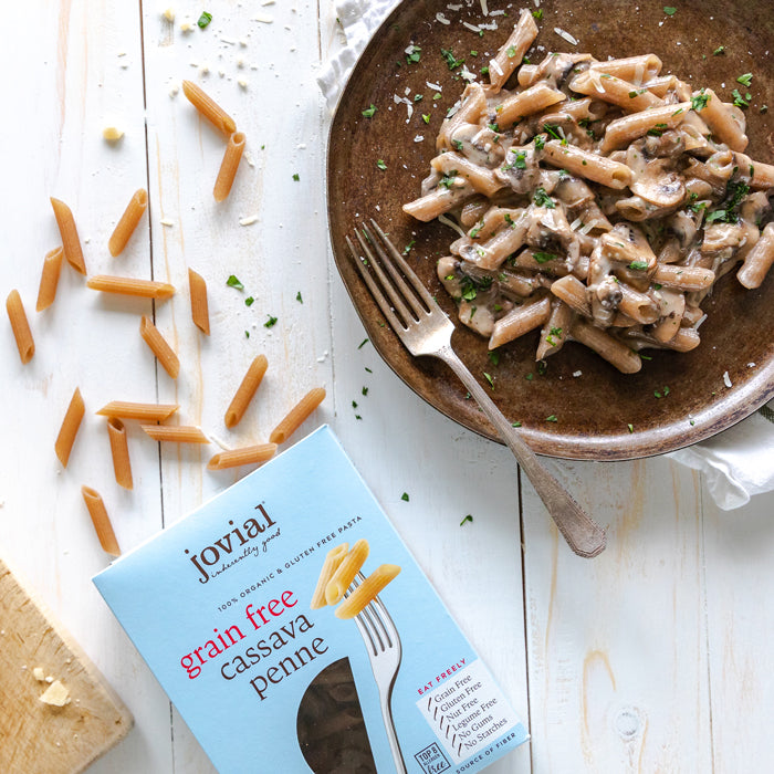 Box of Jovial grain-free cassava penne beside a bowl of creamy mushroom penne with a fork and scattered dry pasta on a white wood surface