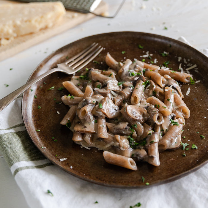 Grain-free penne in creamy mushroom sauce on a brown plate, garnished with parsley and grated cheese, fork at side