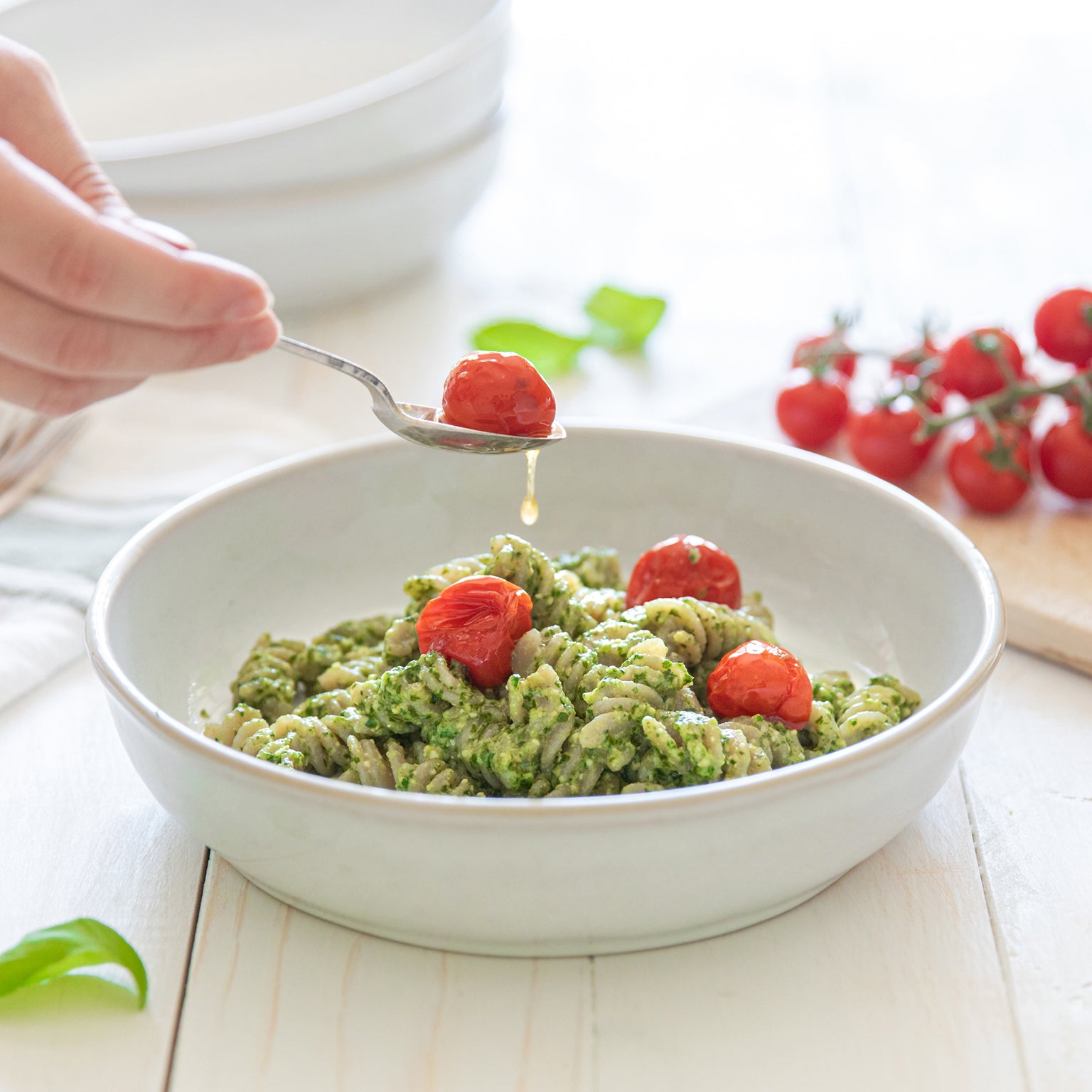 Bowl of cassava fusilli tossed in cashew pesto with tomato confit as a spoon drizzles oil over the pasta