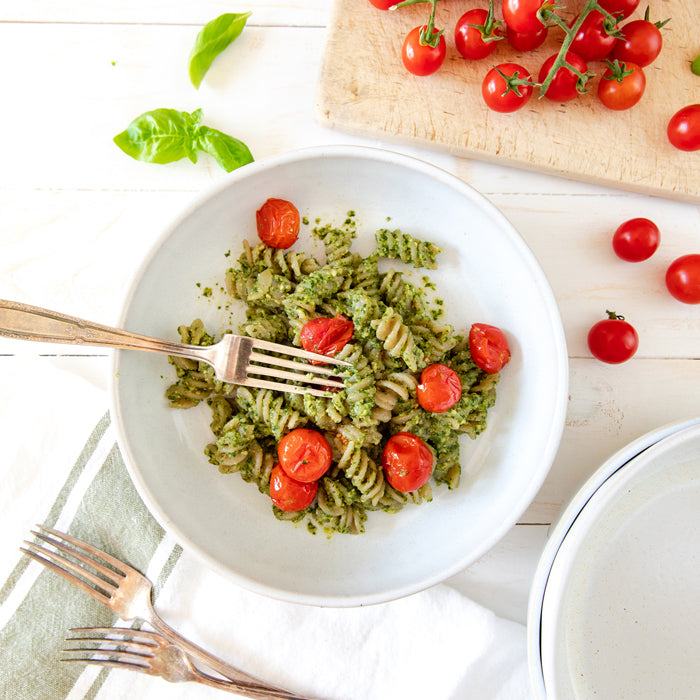 Bowl of cassava fusilli tossed in cashew pesto topped with roasted cherry tomato confit on a white tabletop, fork resting in the pasta