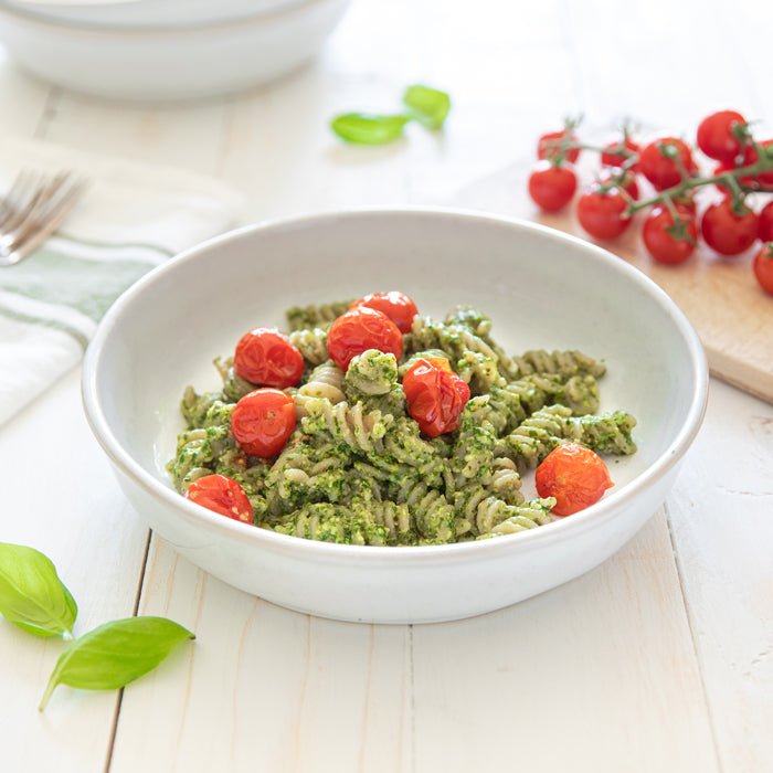Bowl of cassava fusilli pasta tossed in cashew basil pesto, topped with roasted cherry tomatoes on a white wooden table