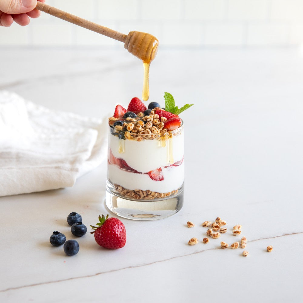 Parfait with yogurt, strawberries, and Organic Einkorn Cereal Puffs topped with granola and honey on a white surface