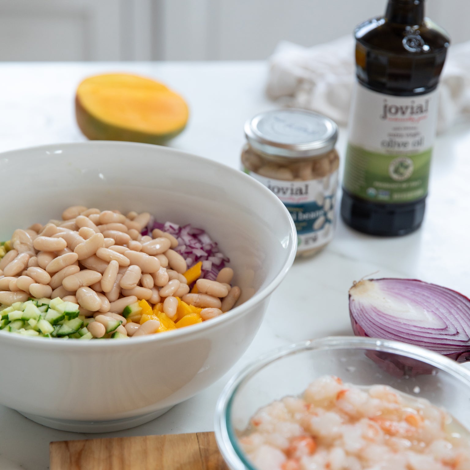 Bowl of cannellini beans, diced mango, cucumber, red onion, and shrimp for ceviche with Jovial olive oil bottle visible